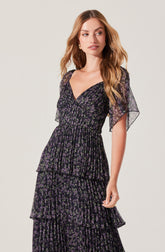 A woman is wearing a black dress with a purple floral pattern. The dress features a deep V neckline, short flutter sleeves, and a fitted waist. The skirt has multiple layers of ruffled fabric, creating a tiered effect.